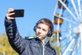 A teenager in a black jacket listens to music on headphones and makes selfie on the background of a Ferris wheel on a sunny autumn Royalty Free Stock Photo