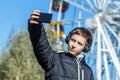 Autumn. A teenager in a black jacket listens to music on headphones and makes selfie on the background of a Ferris wheel on a sunn Royalty Free Stock Photo