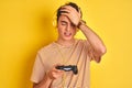 Teenager boy playing video games using gamepad over isolated yellow background stressed with hand on head, shocked with shame and