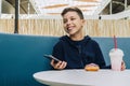 Teenager boy sits at table in cafe, drinks milkshake, eats donut, holds smartphone in his hand. Boy plays mobile games. Royalty Free Stock Photo