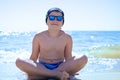 A teenager boy sitting on the sand in swimming trunks, blue sun glasses, is engaged in meditation, relaxation and yoga on the sea Royalty Free Stock Photo