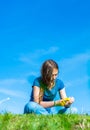 Teenager brunette girl with long hair sit on the grass and wreathes a wreath of yellow dandelion flowers on sky background with co