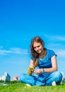 Teenager brunette girl with long hair sit on the grass and wreathes a wreath of yellow dandelion flowers on sky background with co Royalty Free Stock Photo