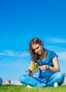Teenager brunette girl with long hair sit on the grass and wreathes a wreath of yellow dandelion flowers on sky background with co
