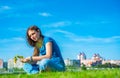 Teenager brunette girl with long hair sit on the grass and wreathes a wreath of yellow dandelion flowers on sky background with co