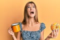 Teenager caucasian girl eating doughnut and drinking coffee angry and mad screaming frustrated and furious, shouting with anger Royalty Free Stock Photo