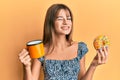 Teenager caucasian girl eating doughnut and drinking coffee winking looking at the camera with expression, cheerful and happy Royalty Free Stock Photo