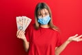 Teenager caucasian girl wearing medical mask holding 10 united kingdom pounds banknotes celebrating achievement with happy smile