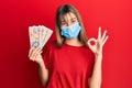 Teenager caucasian girl wearing medical mask holding 10 united kingdom pounds banknotes doing ok sign with fingers, smiling