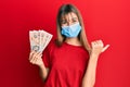 Teenager caucasian girl wearing medical mask holding 10 united kingdom pounds banknotes pointing thumb up to the side smiling