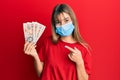 Teenager caucasian girl wearing medical mask holding 10 united kingdom pounds banknotes smiling happy pointing with hand and