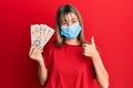 Teenager caucasian girl wearing medical mask holding 10 united kingdom pounds banknotes smiling happy and positive, thumb up doing