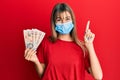 Teenager caucasian girl wearing medical mask holding 10 united kingdom pounds banknotes smiling with an idea or question pointing