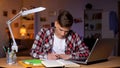 Teenager doing his homework, writing task into copybook, laptop on table