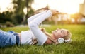 Teenager girl lies in grass in a park and listening to music on headphones while holding her smartphone overhead and typing on the Royalty Free Stock Photo