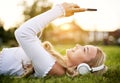 Teenager girl lies in grass in a park and listening to music on headphones while holding her smartphone overhead and typing on the Royalty Free Stock Photo