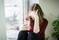 A teenager girl with long blond hair sits by the window on a windowsill with a phone in her hand and covered her face with her