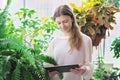 teenager girl looking at pc tablet standing among large geen plants. home jungle concept. looking for plant care tips