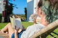 Teenager girl resting lying in an outdoor chair on lawn, reading book, drinks water from bottle Royalty Free Stock Photo