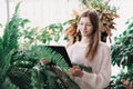 teenager girl standing between large green plants and looking at pc tablet. home jungle concept. teen girl looking for