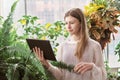 teenager girl standing between large green plants and looking at pc tablet. home jungle concept. teen girl looking for
