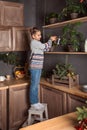 Teenager girl in a sweater and jeans stands on a stool and watering flowers on the upper shelves of the kitchen in the evening