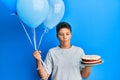 Teenager hispanic boy celebrating birthday with cake holding balloons making fish face with mouth and squinting eyes, crazy and Royalty Free Stock Photo