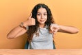 Teenager hispanic girl wearing casual clothes sitting on the table doing thumbs up and down, disagreement and agreement expression Royalty Free Stock Photo