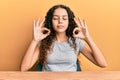 Teenager hispanic girl wearing casual clothes sitting on the table relaxed and smiling with eyes closed doing meditation gesture Royalty Free Stock Photo