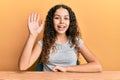 Teenager hispanic girl wearing casual clothes sitting on the table waiving saying hello happy and smiling, friendly welcome