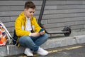 Teenager with mobile phone sitting next to his electric scooter and backpack. Modern young boy chatting on smartphone. Royalty Free Stock Photo