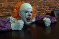 A teenager in a scary clown costume with sharp teeth sits at a cafe table and clenched his hands into fists. Street makeup on a Royalty Free Stock Photo