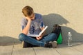 Teenager schoolboy reads textbook, sits near the gray wall of the school. Royalty Free Stock Photo