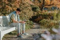 A Teenager Sits On A Bench In The Autumn Park Drinks Coffee From A Thermo Mug And Looks Into A Phone. Portrait Of