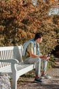 A Teenager Sits On A Bench In The Autumn Park Drinks Coffee From A Thermo Mug And Looks Into A Phone. Portrait Of