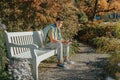 A Teenager Sits On A Bench In The Autumn Park Drinks Coffee From A Thermo Mug And Looks Into A Phone. Portrait Of