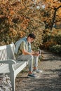 A Teenager Sits On A Bench In The Autumn Park Drinks Coffee From A Thermo Mug And Looks Into A Phone. Portrait Of