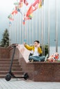 Teenager sits near the administration building against background of country flags and waves into the smartphone. Electric scooter