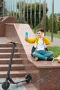 Teenager sits near the administration building against background of country flags and waves into the smartphone. Electric scooter