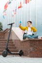 Teenager sits near the administration building against background of country flags and waves into the smartphone. Electric scooter