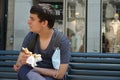 A teenager sitting on a bank in ZÃÂ¼rich in corona time, eating a butter bretzel, his facial mask is attached to his rucksack