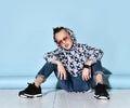 Teenager in sunglasses, blue jeans, hoodie, black bracelet and sneakers. Raised hand up, sitting on white floor. Blue background Royalty Free Stock Photo