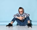 Teenager in sunglasses, blue jeans, hoodie, black bracelet and sneakers. Raised hand up, sitting on white floor. Blue background Royalty Free Stock Photo