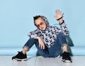 Teenager in sunglasses, blue jeans, hoodie, black bracelet and sneakers. Raised hand up, sitting on white floor. Blue background Royalty Free Stock Photo