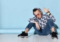 Teenager in sunglasses, blue jeans, hoodie, black bracelet and sneakers. Raised hand up, sitting on white floor. Blue background Royalty Free Stock Photo