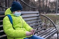 A teenager wearing a medical mask on a Park bench uses a smartphone and headphones. Royalty Free Stock Photo