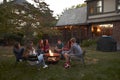 Teenagers sit talking around a fire pit in a garden at dusk