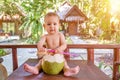 Teething and salivation infant baby at tropical vacation. Eats and drinks green young coconut. Sits on a table Royalty Free Stock Photo