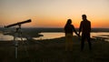 Telescope stands in front of couple holding hands on the hill at the amazing sunset