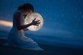 Tender image of a girl; female magic. Beautiful attractive girl on a night beach with sand and stars hugs the moon, art photo. On Royalty Free Stock Photo